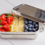 Stainless steel lunch box with steel lid and latching clips, filled with strawberries, banana slices, and blueberries (illustrative).